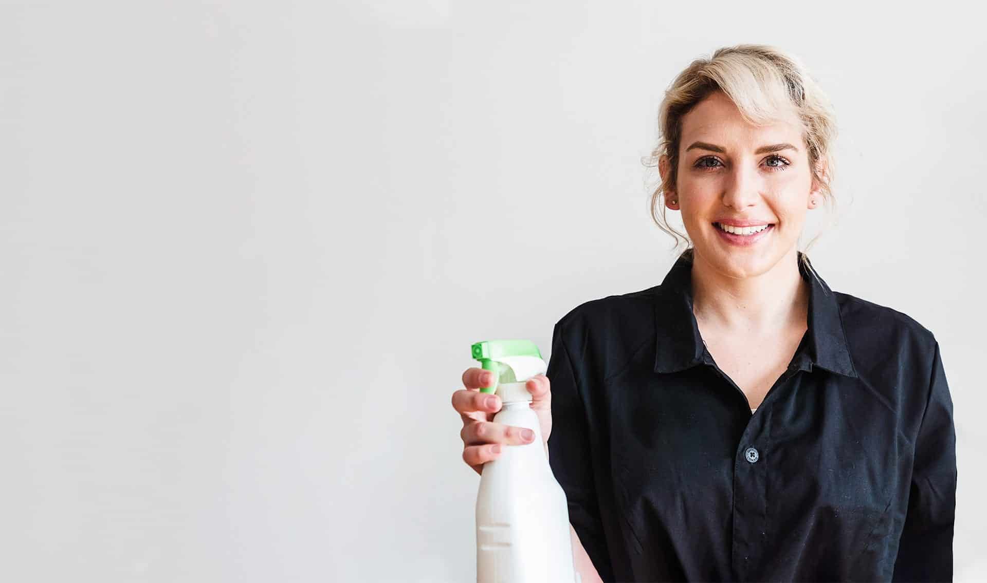 Putzhilfe Darmstadt Blonde mittelalte Frau mit Zopf im dunklen Shirt hält eine Sprühflasche in der Hand und lächelt frontal in die Kamera